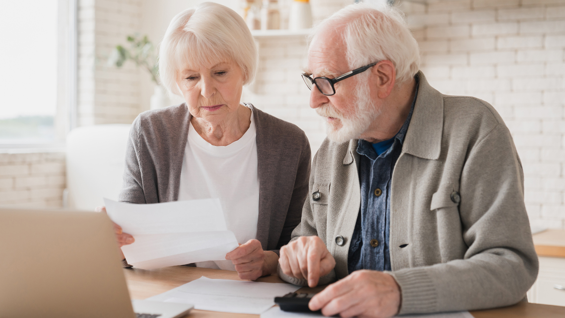 Senior couple counting funds on calculator, at home using laptop.